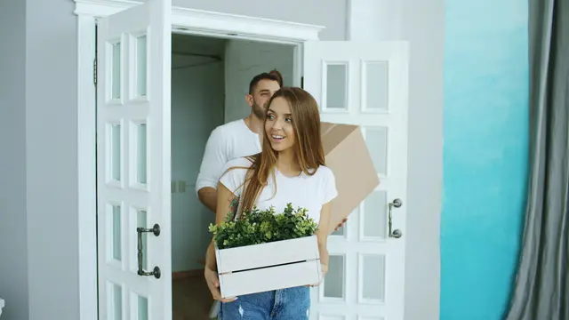 A couple carrying a plant and a box into their new home